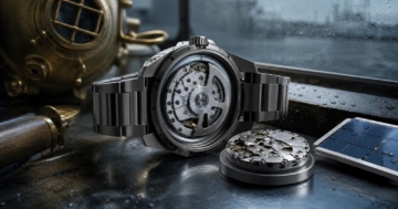 Eine offene Uhr mit titanfarbenem Gehäuse liegt auf einem metallenen Werkstatttisch, daneben liegen ein Uhrwerk, ein antiker