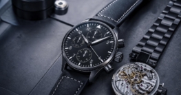 Eine limitierte Jubiläums-Fliegeruhr liegt neben einem offenen Uhrwerk und einem Armband auf dunklem Metall.
