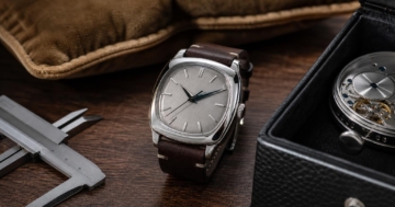 Eine silberne kissenförmige Armbanduhr mit brauntem Lederband liegt auf einer Holzplatte neben einem Messschieber und einer