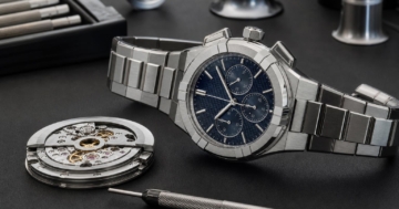 Ein polierter AIKON-Chronograph liegt auf einer dunklen Designer-Schreibtischplatte neben einem losen Uhrwerk und einem