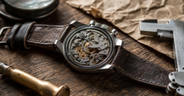 Eine geöffnete Vintage-Chronograph-Uhr mit sichtbarem Uhrwerk liegt auf einem Holztisch neben Werkzeugen und einem