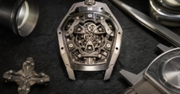 Geöffnete Titan-Uhr mit sichtbarer T-Rahmen-Kinematik, vier dreiseitigen Prismen und Malta-Kreuz-Rad auf dunklem